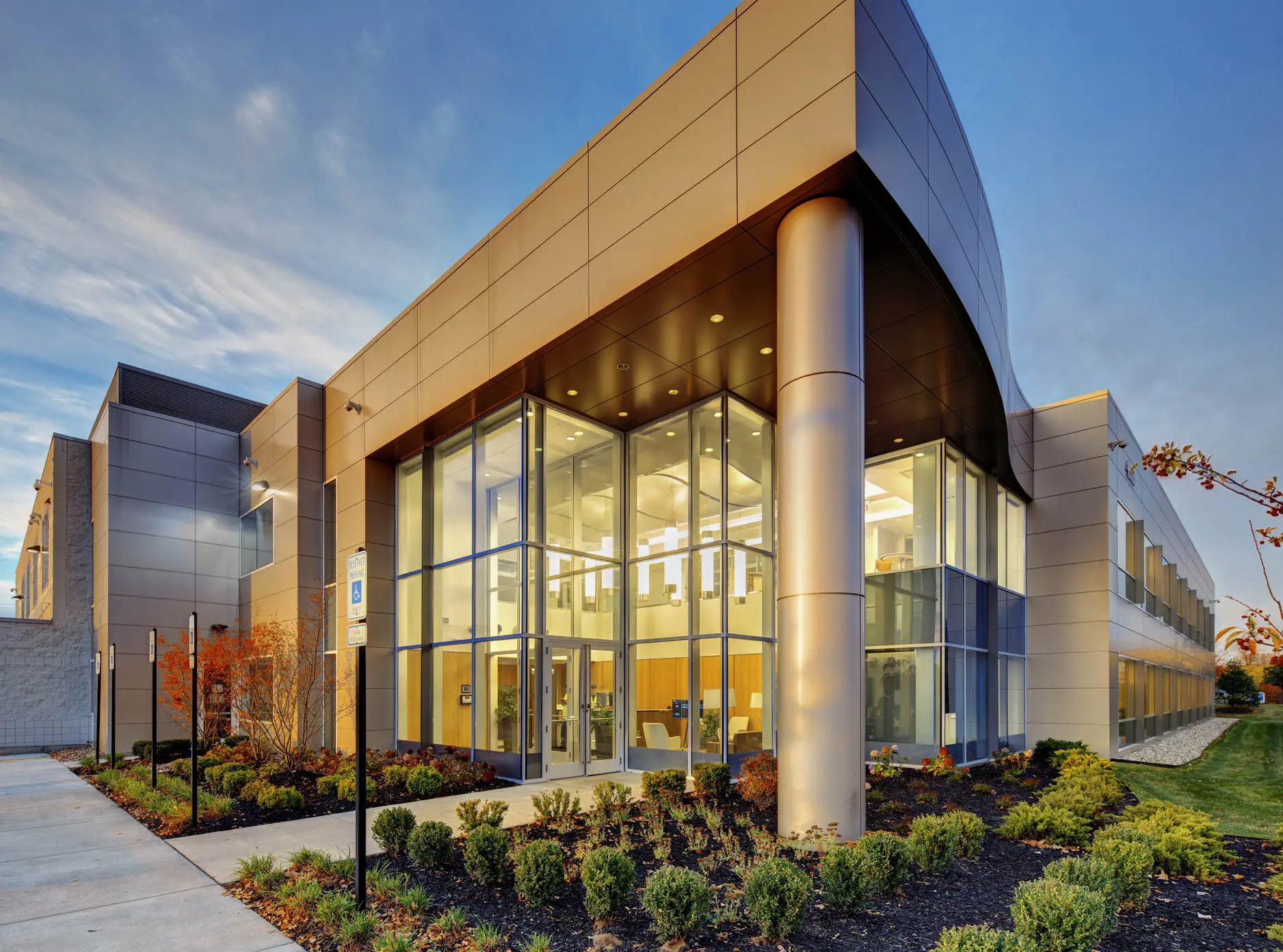 Modern commercial building entrance with curved metal canopy, glass walls and landscaped grounds.