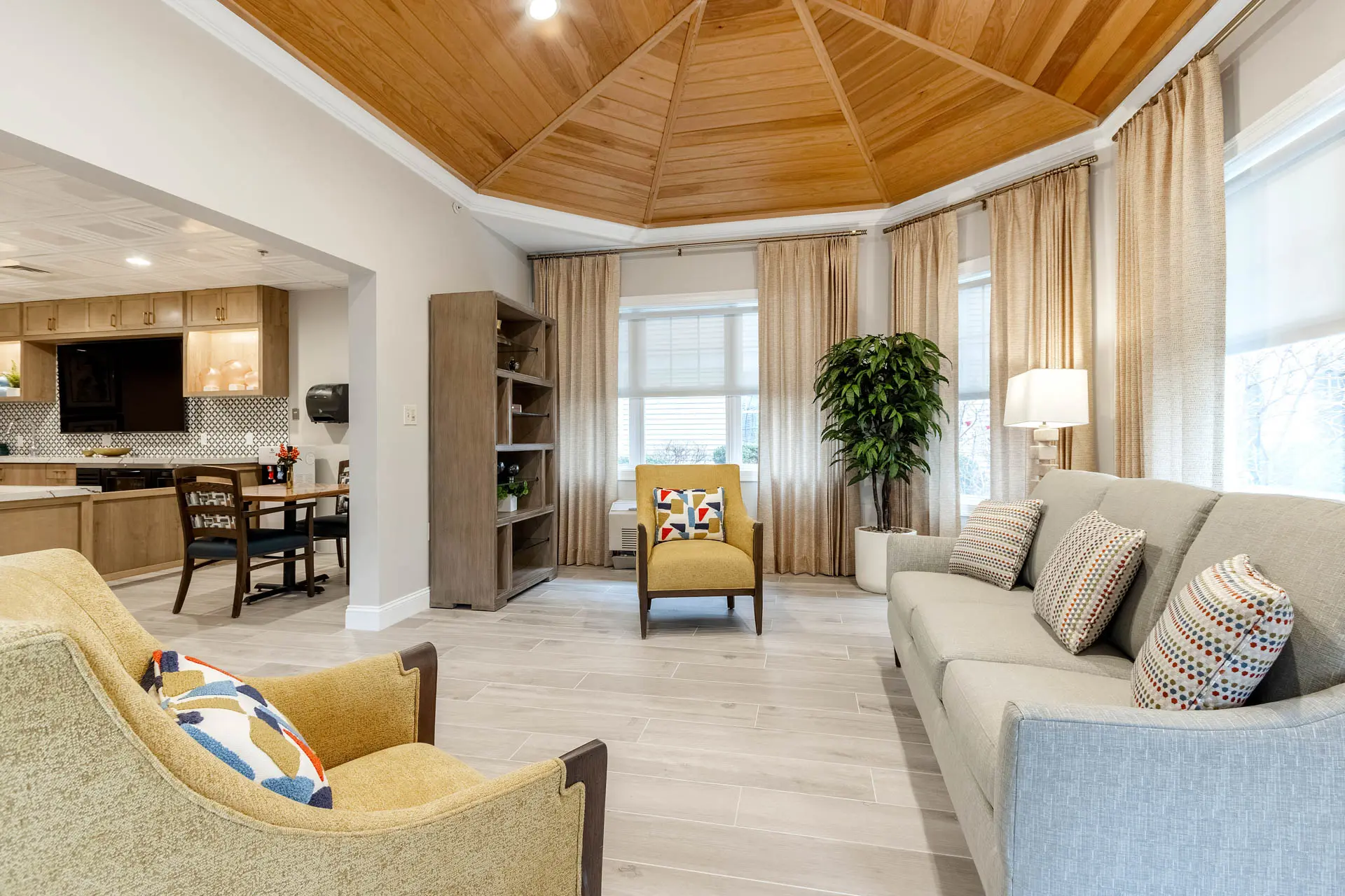 Bright senior living sunroom with sofa, armchairs, wood ceiling and large windows.